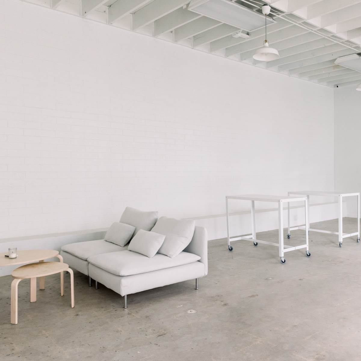 A minimalistic interior space with a light gray sofa, two wheeled white tables, and a large mirror against a white brick wall.