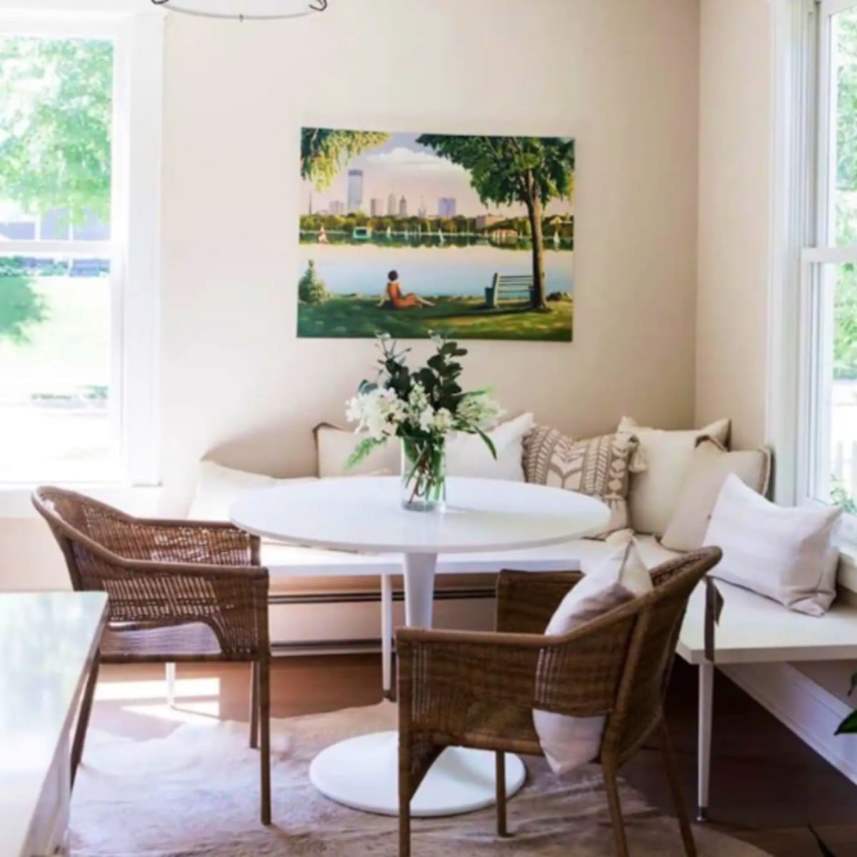 A dining area features a round white table surrounded by wicker chairs and adorned with a vase of flowers, with a large painting of a park scene on the wall.