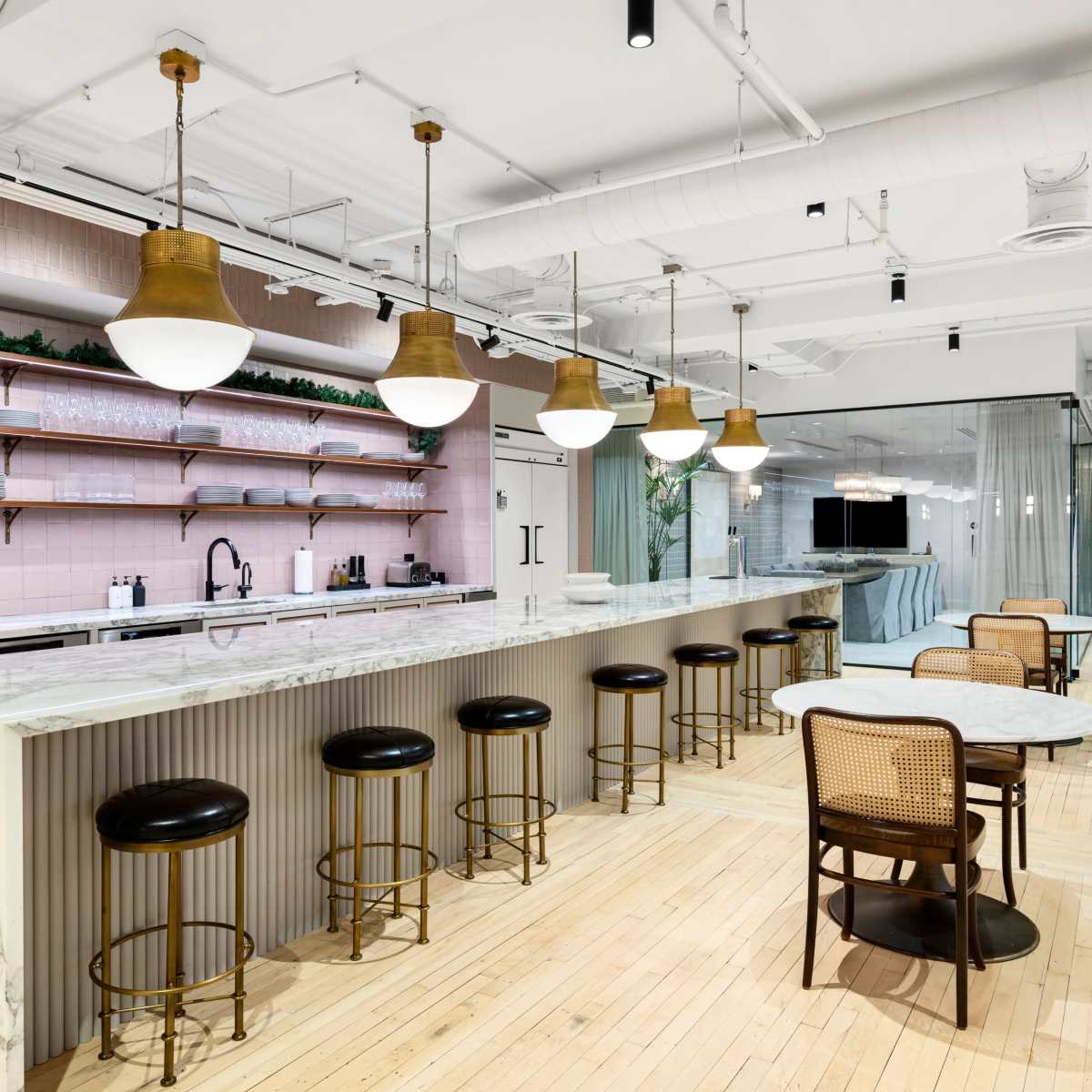 A modern kitchen area with a long marble countertop, bar stools, and wooden floors, featuring shelves with greenery and glass paneling in the background.