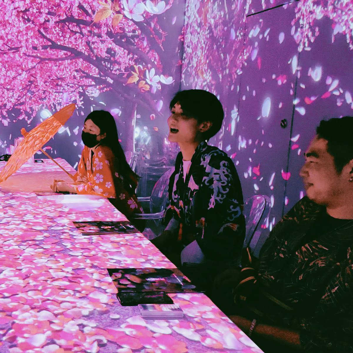 A group of six people sits around a table surrounded by a vibrant floral projection, engaging in conversation.