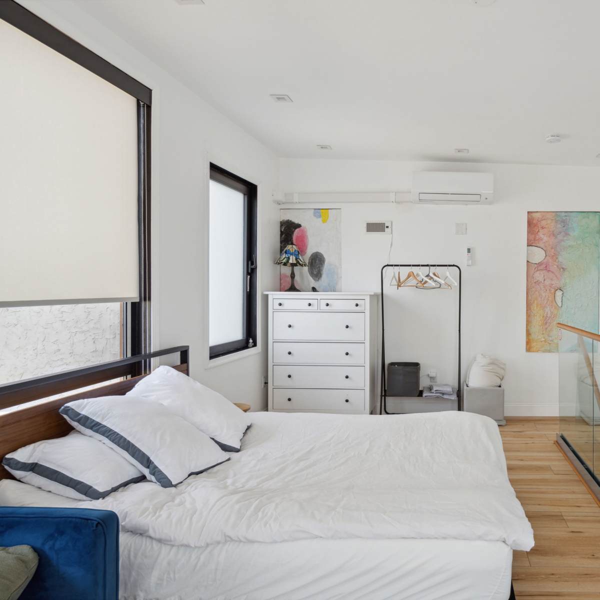 A modern, minimalist bedroom featuring a unmade bed, a dresser, and a clothing rack, with large windows allowing natural light to enter the space.
