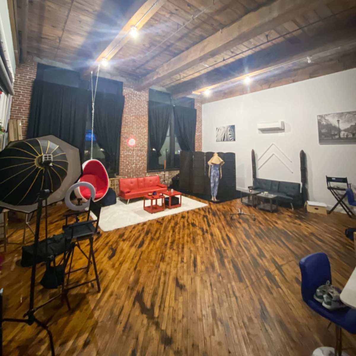 A spacious, well-lit loft featuring wooden floors, a red sofa, various chairs, and photography equipment against exposed brick walls.