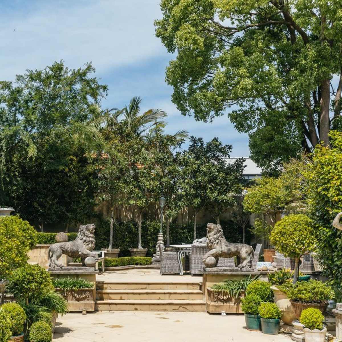 A landscaped garden features stone lion statues flanking a staircase, surrounded by lush greenery and a glass conservatory in the background.