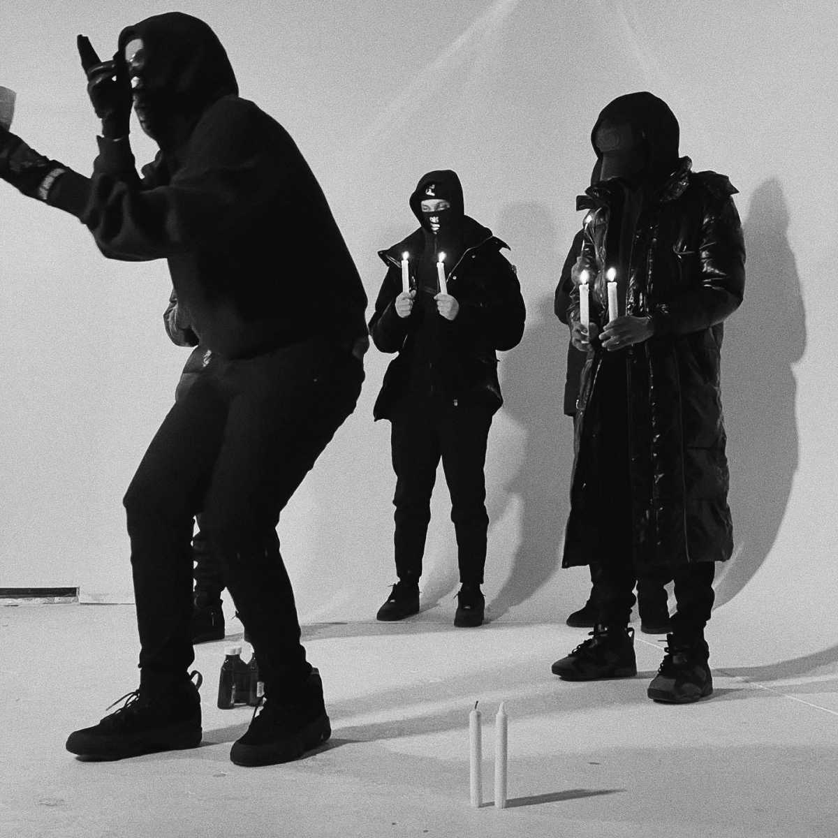 Four individuals in dark clothing and masks hold candles in a dimly lit space.