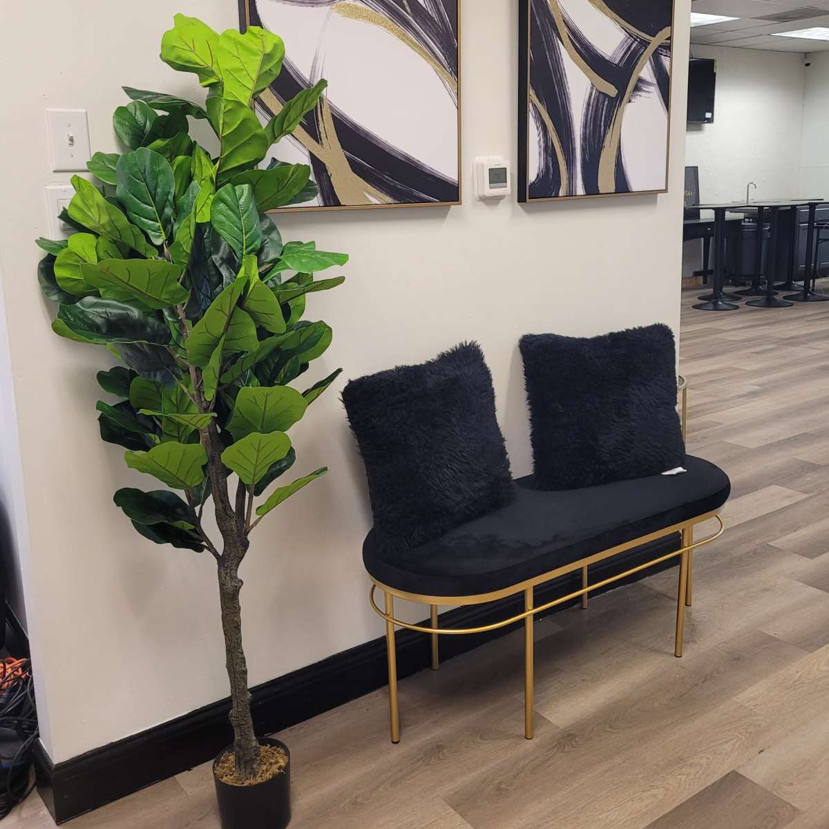 A small seating area features a gold-framed bench with black cushions, complemented by a potted leafy plant and two abstract art pieces on the wall.
