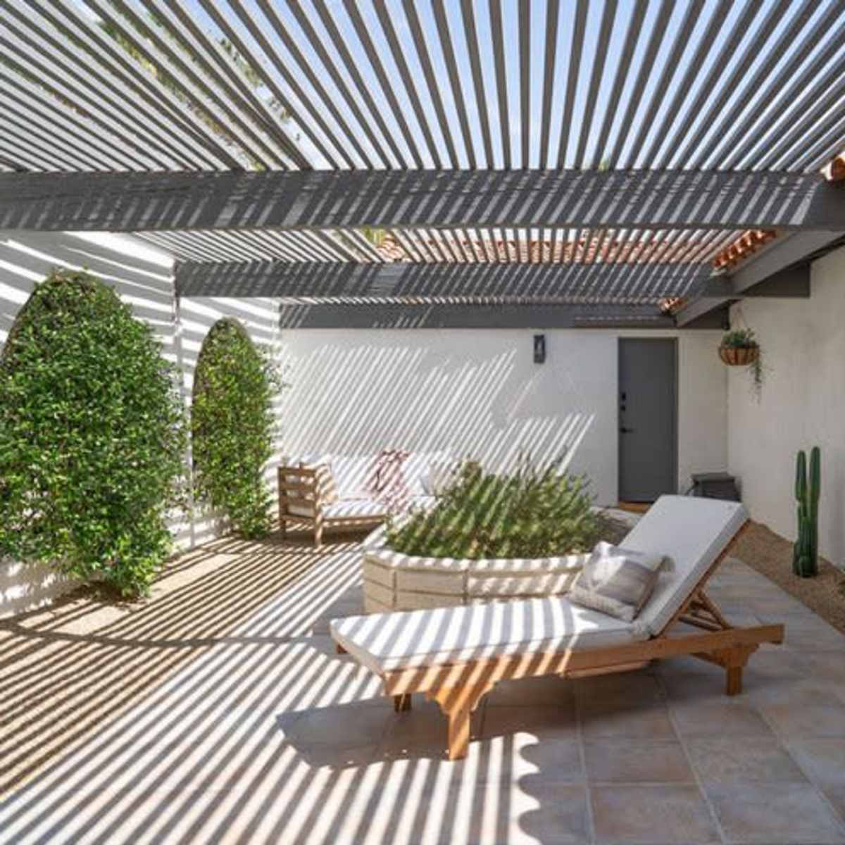 A patio with a lounge chair and a circular seating area, surrounded by potted plants and cast shadows from a slatted roof.