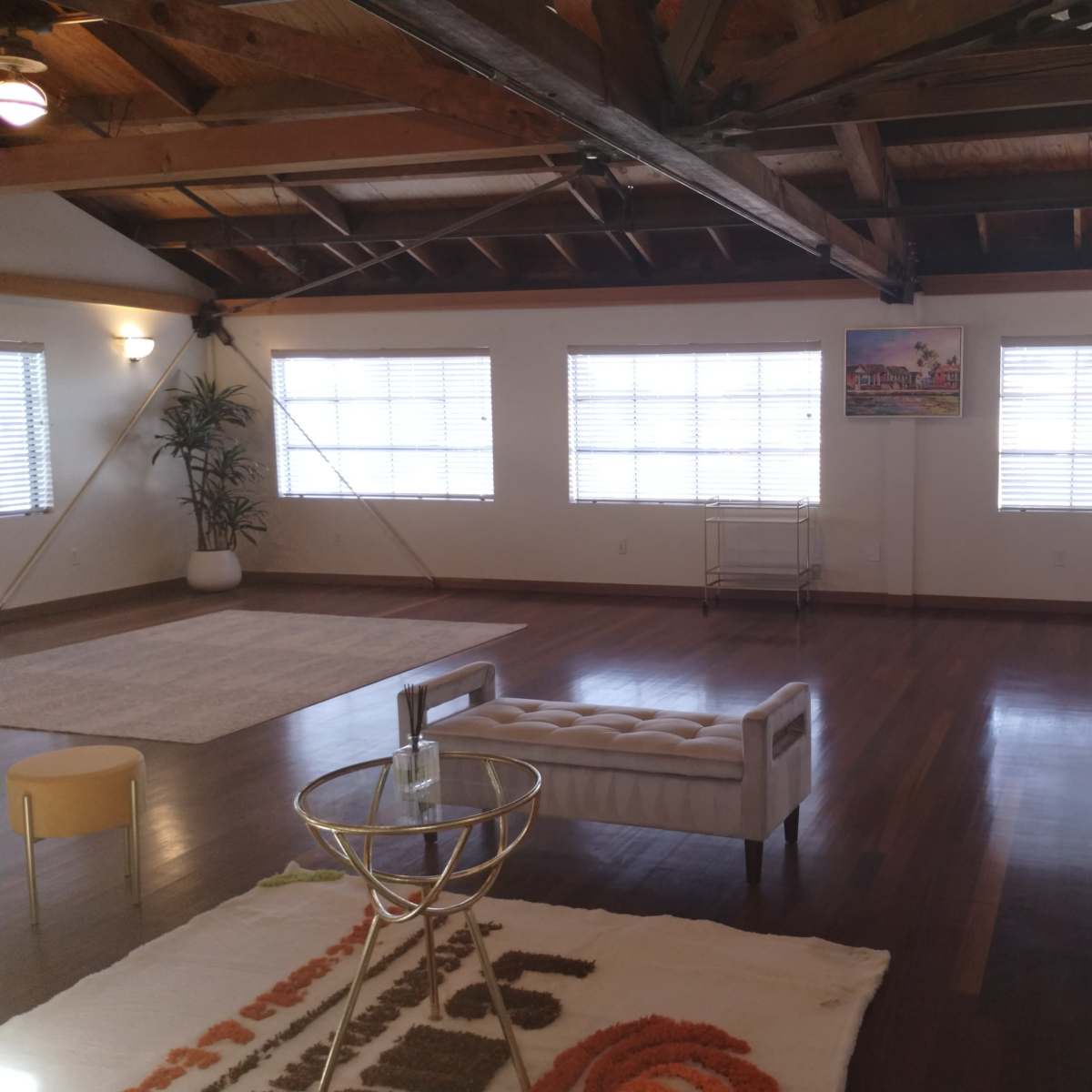A spacious room with wooden beams, large windows covered by blinds, and minimalist furniture including a tufted sofa and a coffee table.