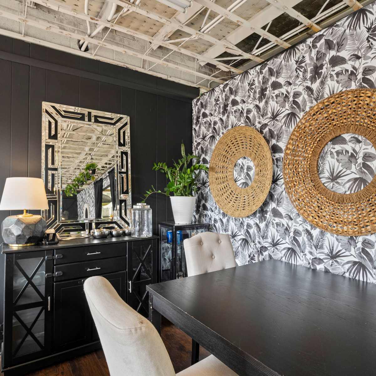 A dining area featuring a black table, light-colored chairs, a cabinet, and a wall adorned with patterned wallpaper and woven round wall decorations.
