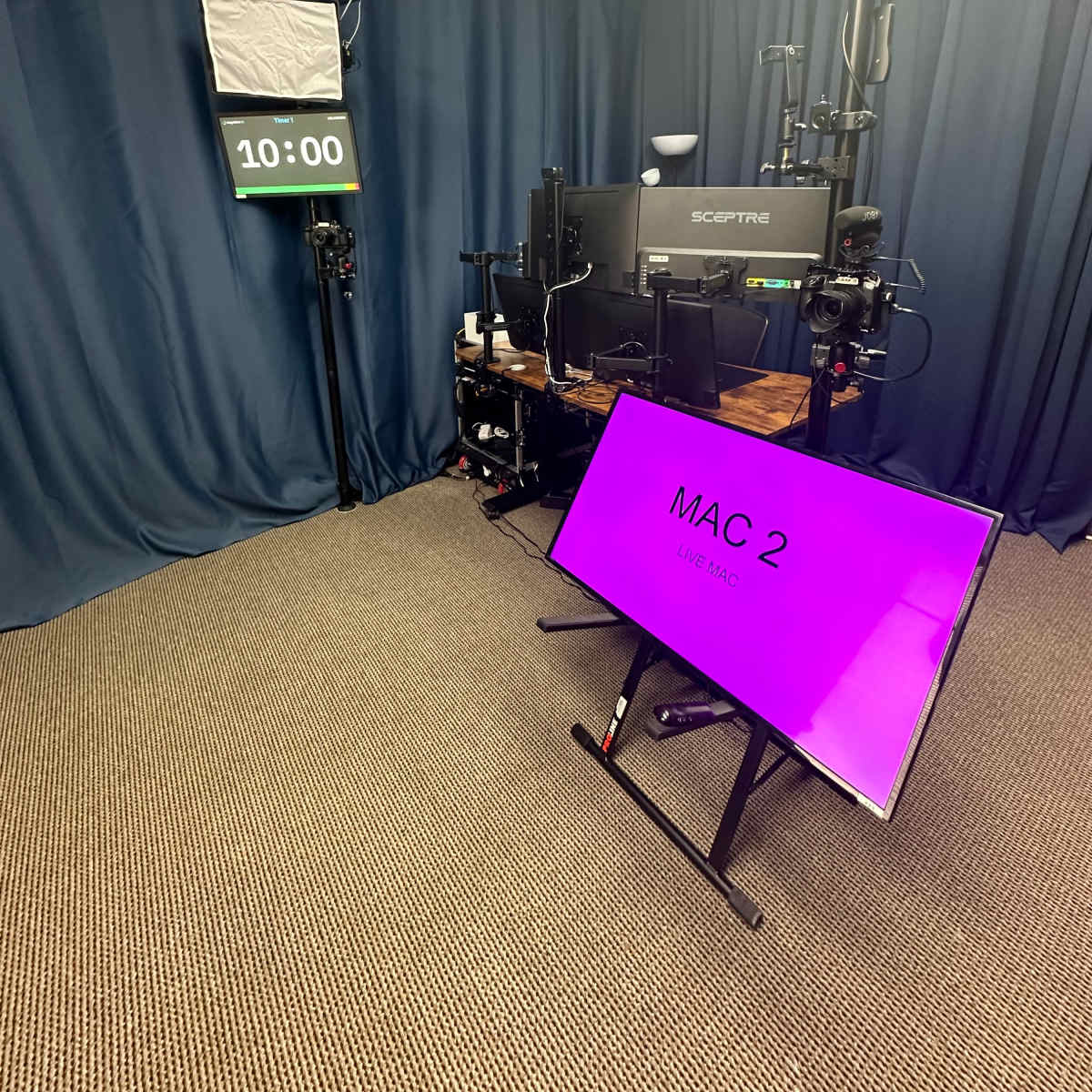 A recording setup featuring a large monitor displaying "MAC 2," a digital clock set to 10:00, and black curtains in the background.