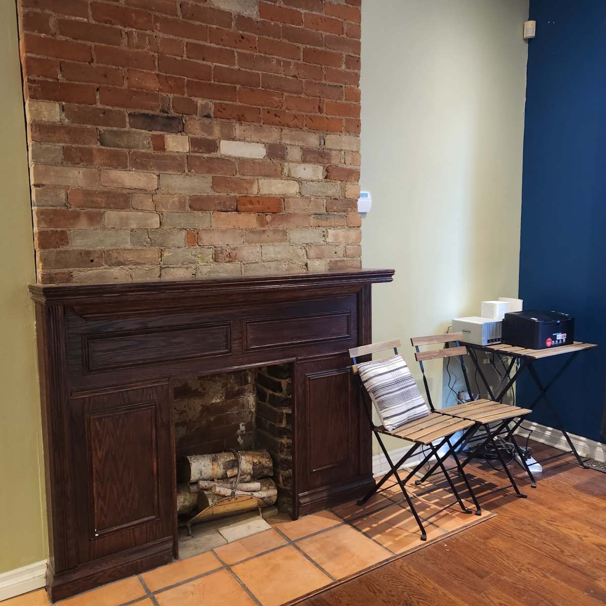 A brick wall with a wooden mantel and an empty fireplace, accompanied by a small wooden table and chair set beside it.