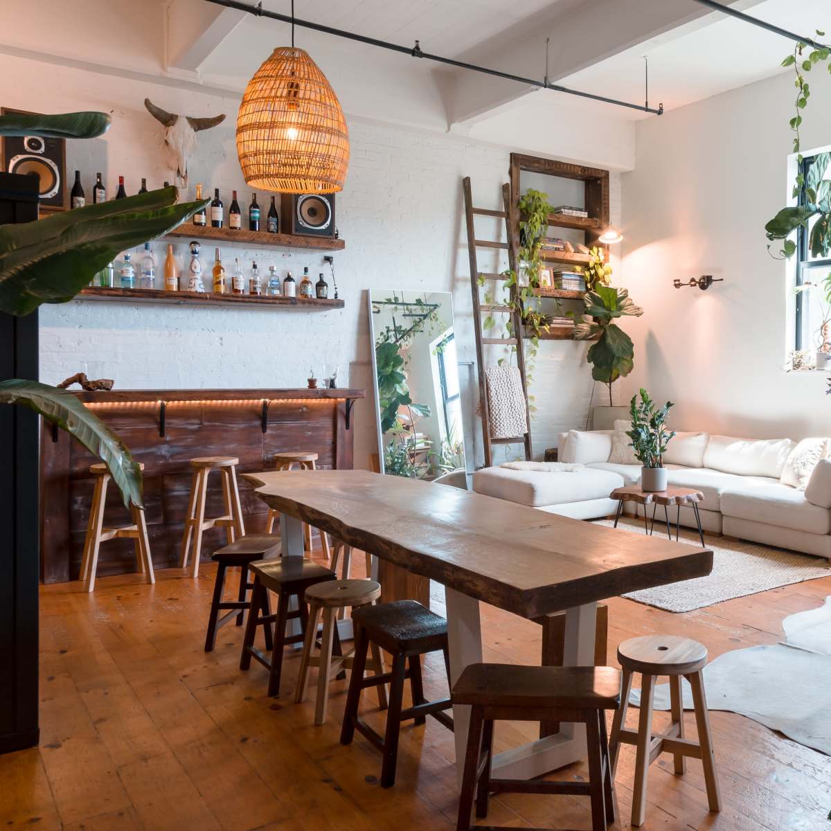A bright, open living space featuring a wooden dining table surrounded by stools, a sofa, and various plants, along with a bar area on one side.