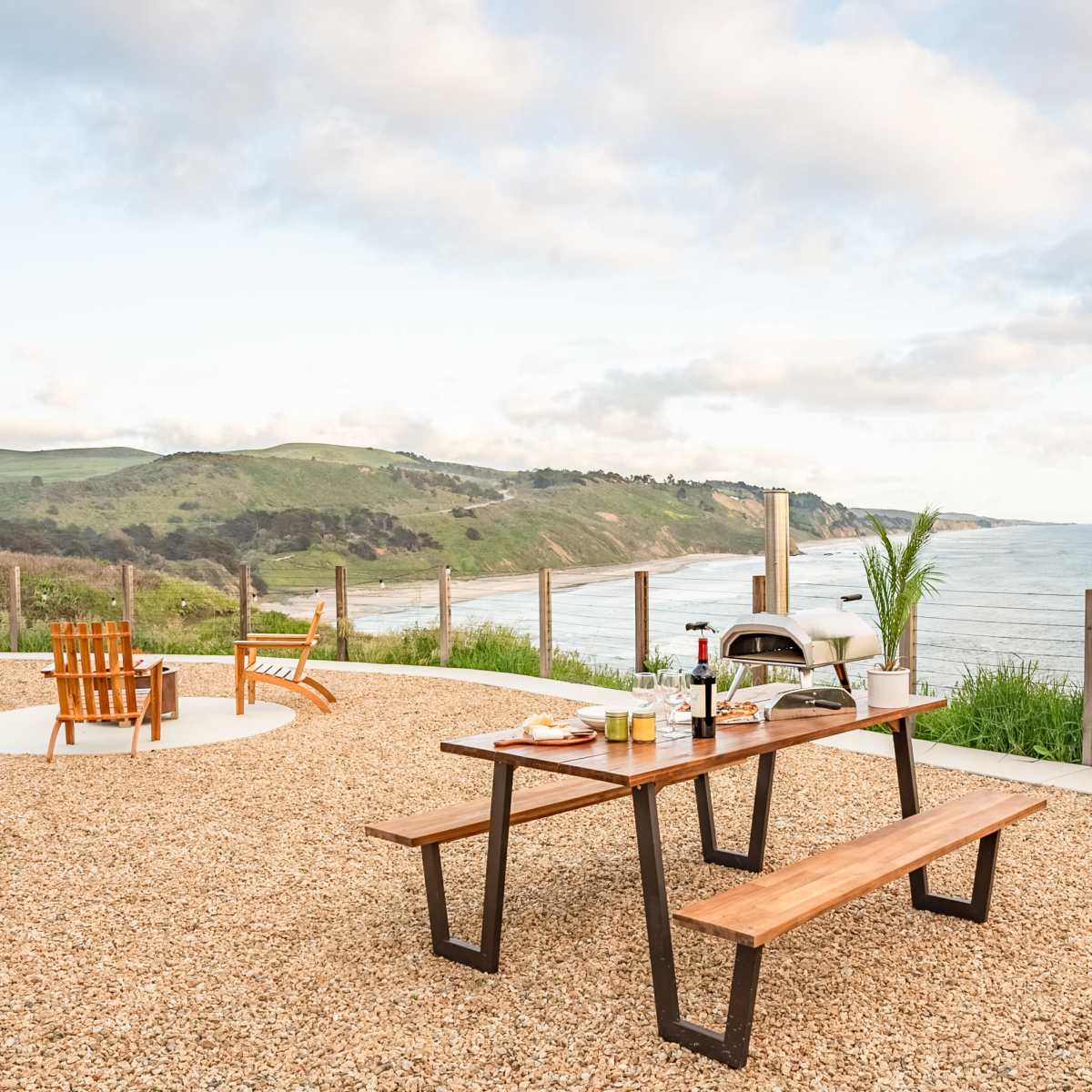 A wooden picnic table with food and drinks is set up in a coastal area overlooking the ocean and hills.
