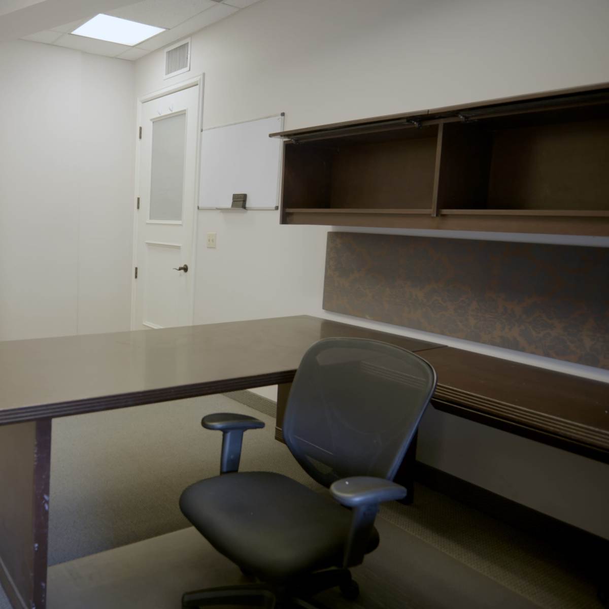 An empty office space features a desk with a swivel chair, a wall-mounted whiteboard, and minimal furnishings.