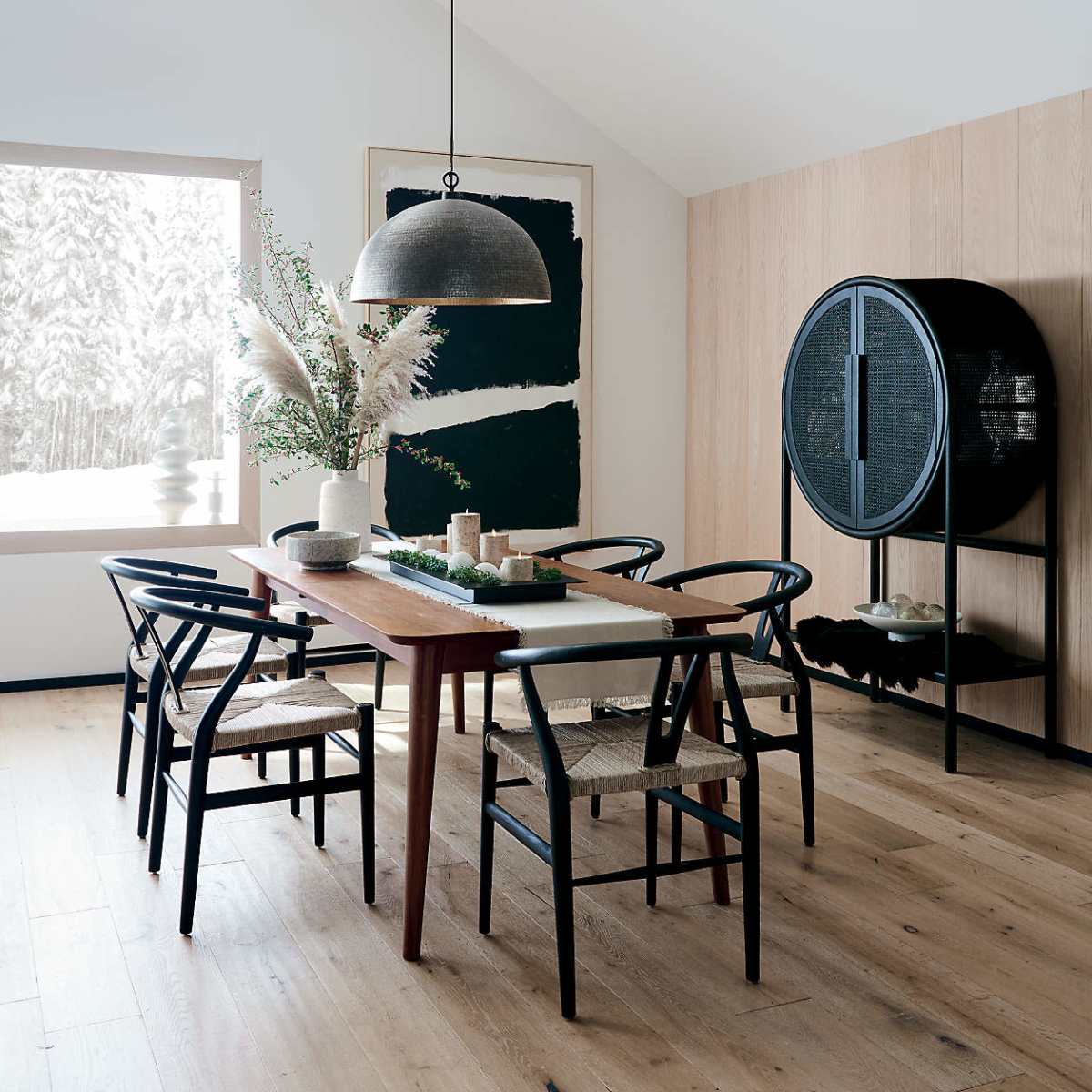 A modern dining room features a wooden table with four black chairs, a large pendant light, and a wall adorned with a painting and decorative elements.