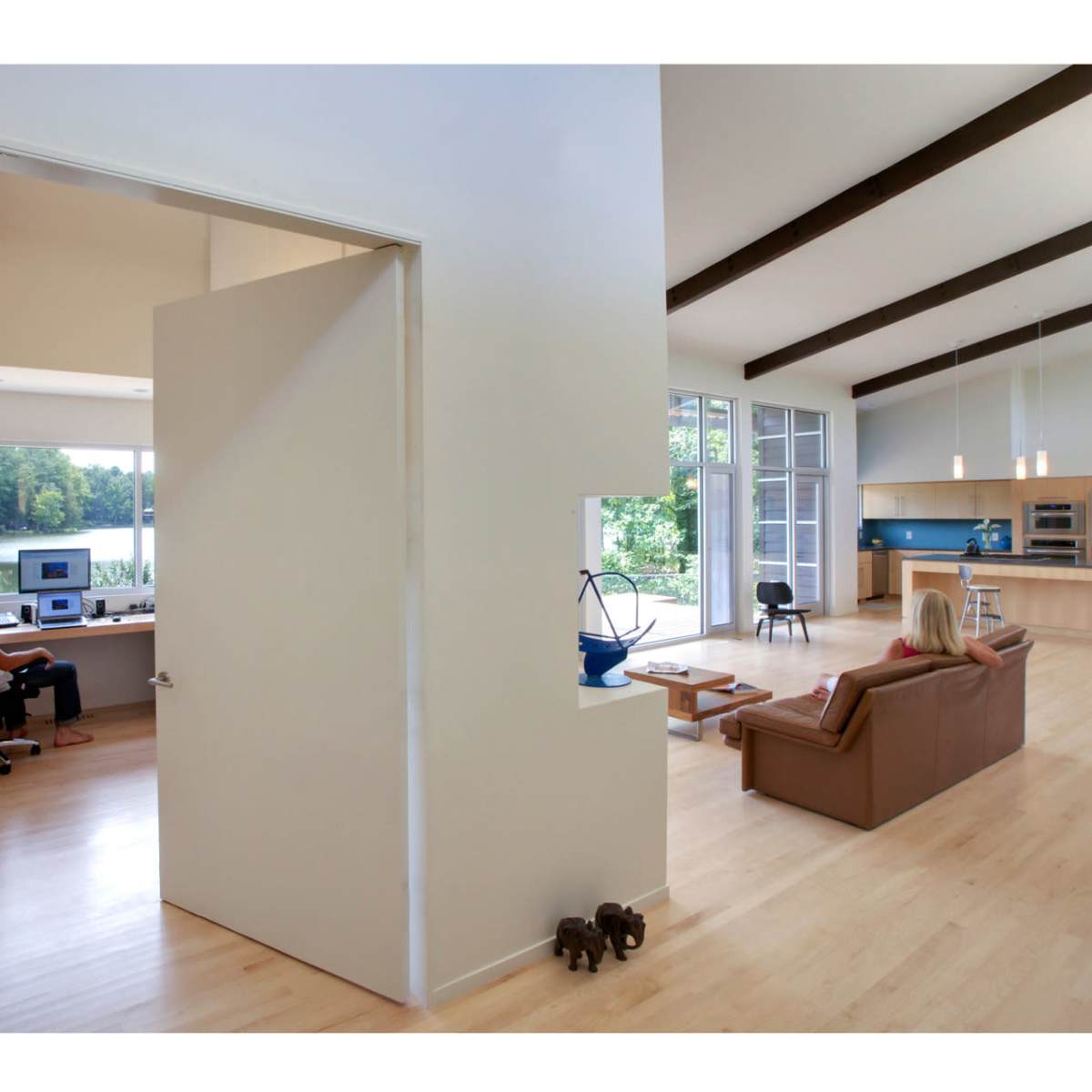 A spacious interior shows a home office on the left with a person working at a desk and a living area on the right overlooking a lake through large windows.