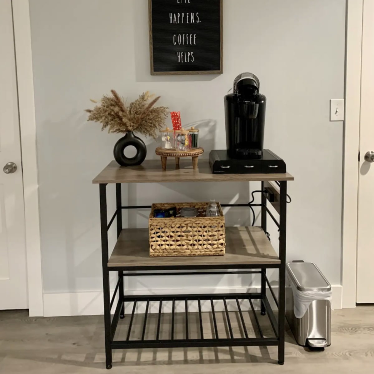 A coffee station with a coffee maker, decorative items, and a basket sits against a gray wall beneath a framed sign that reads "Life happens. Coffee helps."