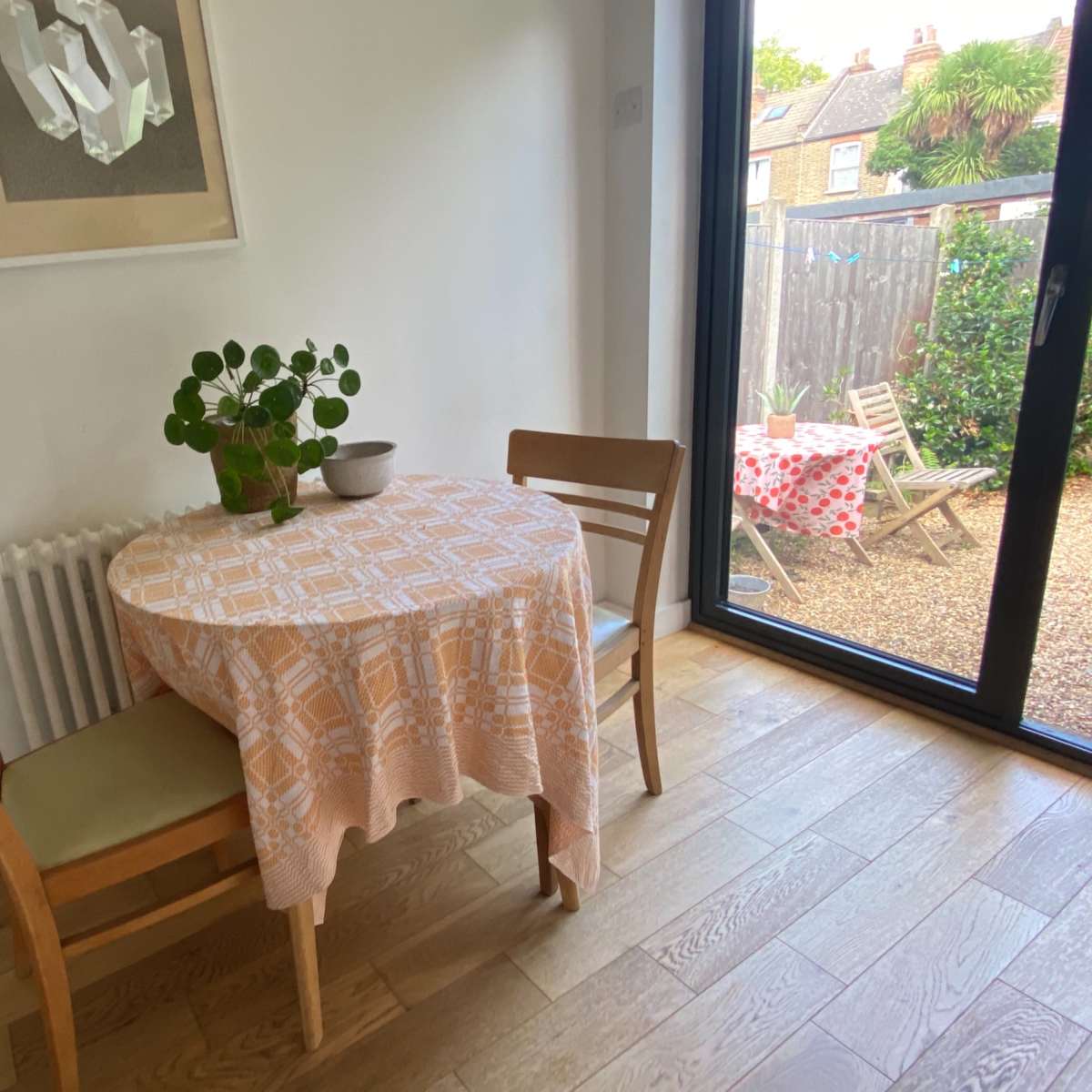 A small dining table with a tablecloth and a plant centerpiece is positioned near a large glass door leading to an outdoor area, with a second table visible in the background.