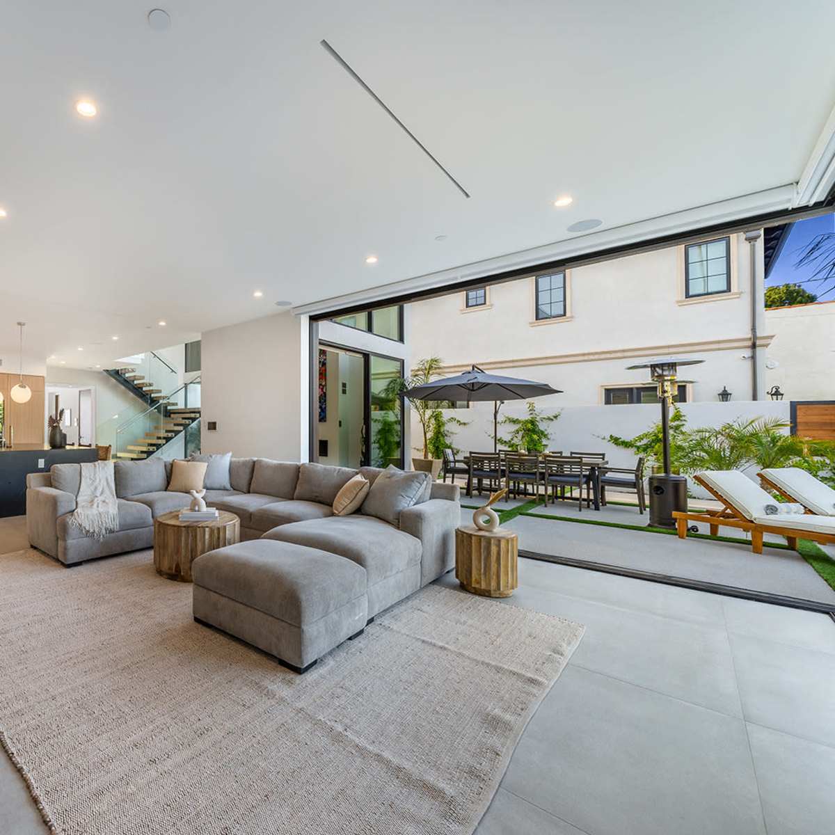 A modern living room with large sliding doors opening to a patio and garden area, featuring a sectional sofa, a coffee table, and a flat-screen TV mounted on the wall.