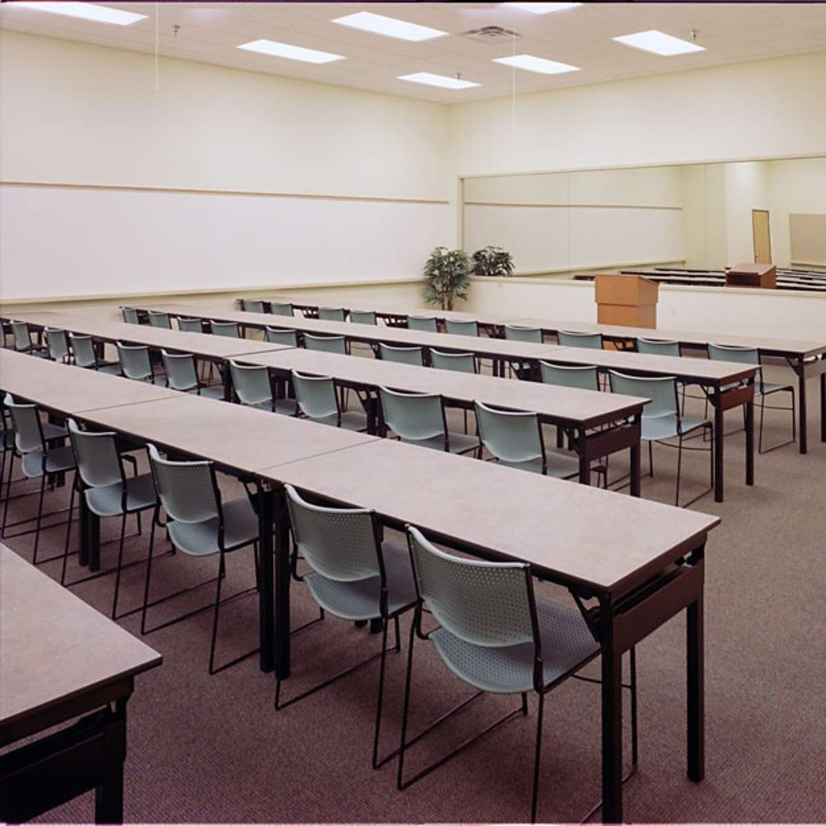 A classroom is set up with rows of tables and chairs facing a wooden podium and a mirrored wall.