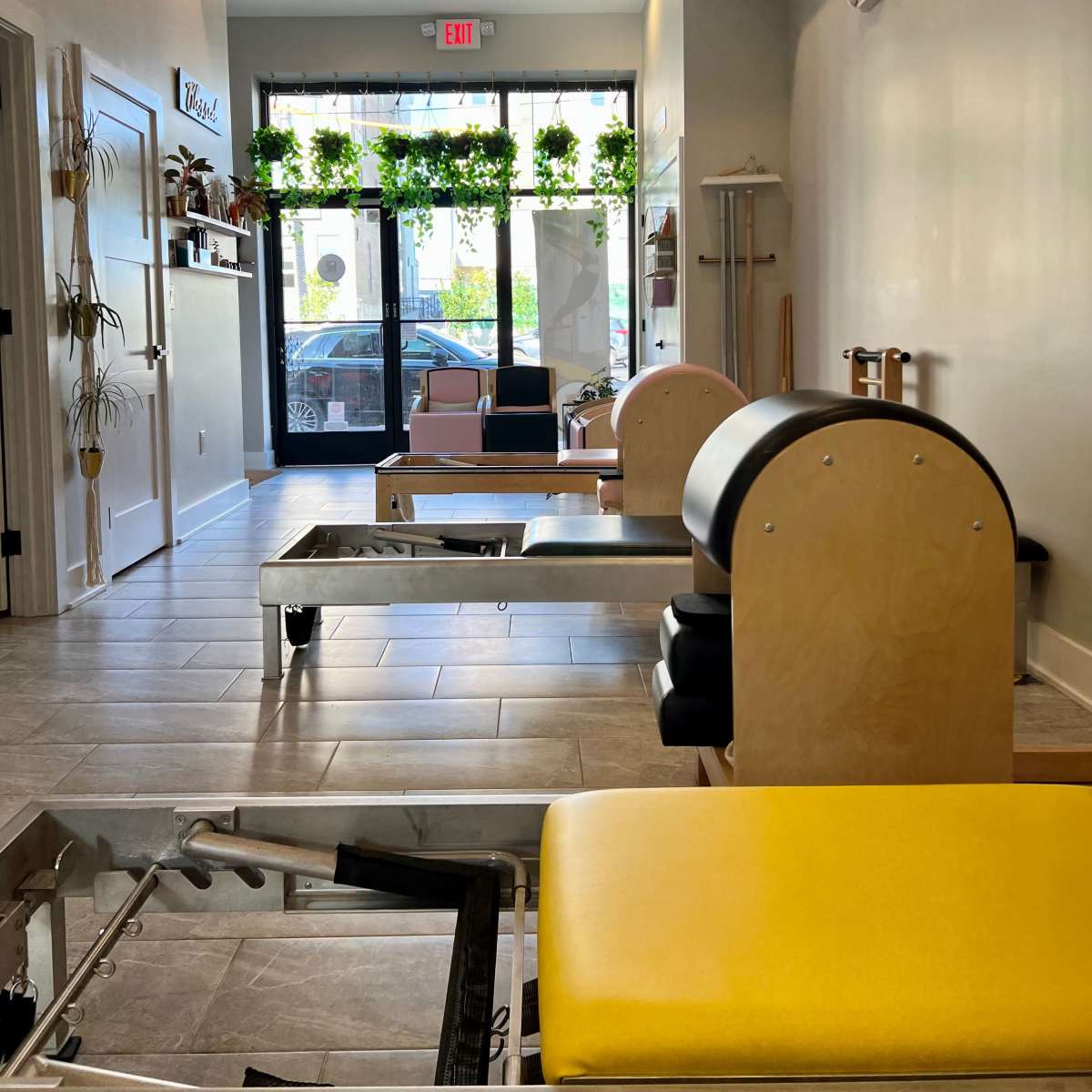 The interior of a studio featuring several Pilates reformers, with large windows displaying plants and natural light.
