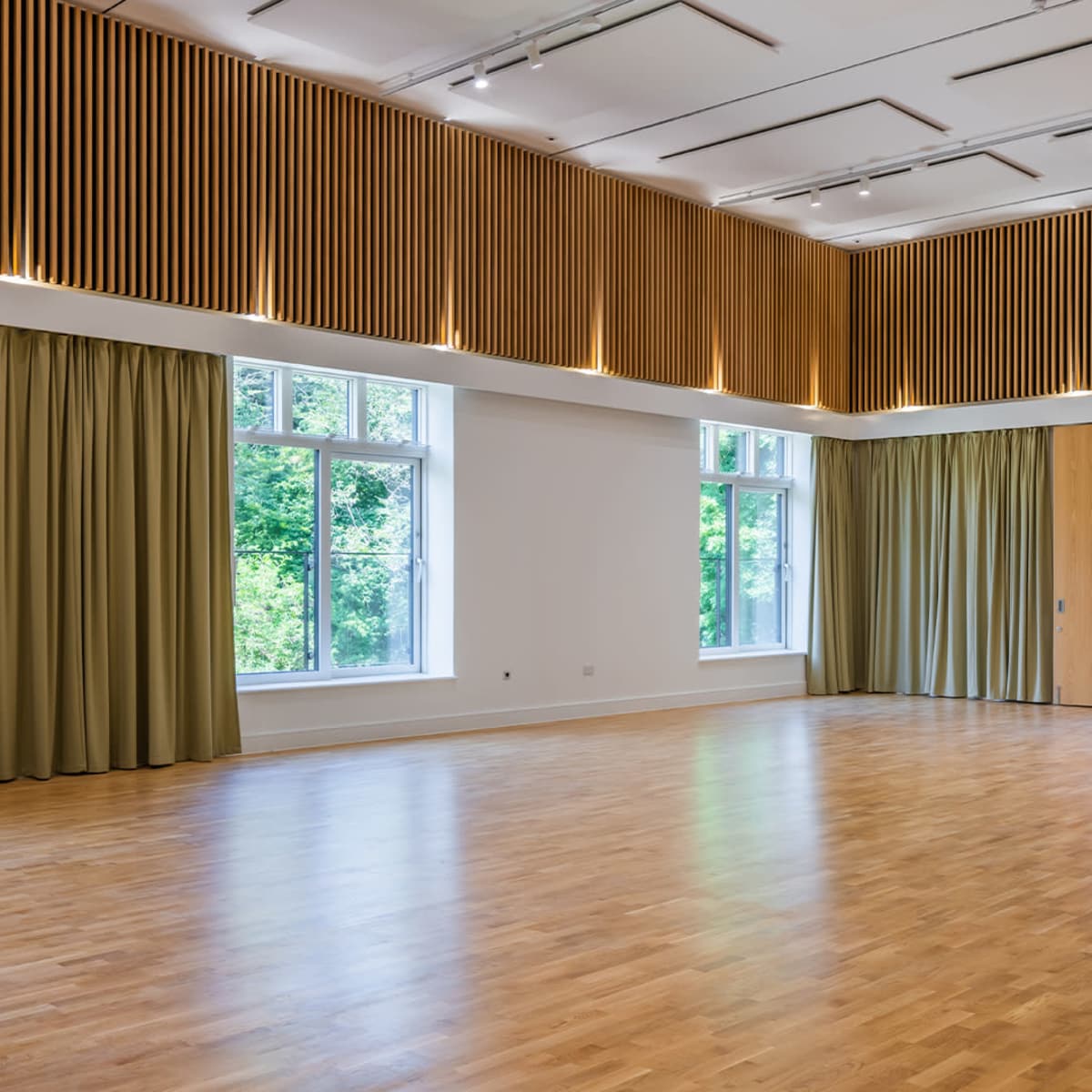 A spacious room with wooden floors, large windows, and curtains, featuring a wood-paneled wall at the top.
