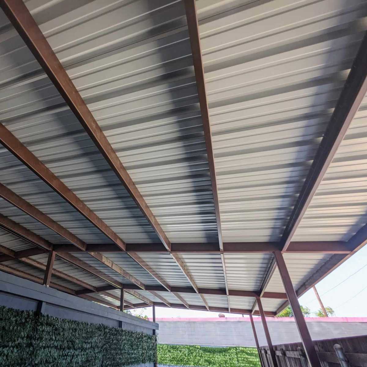 A view of a metal roof's underside supported by steel beams, alongside a wall covered in greenery.