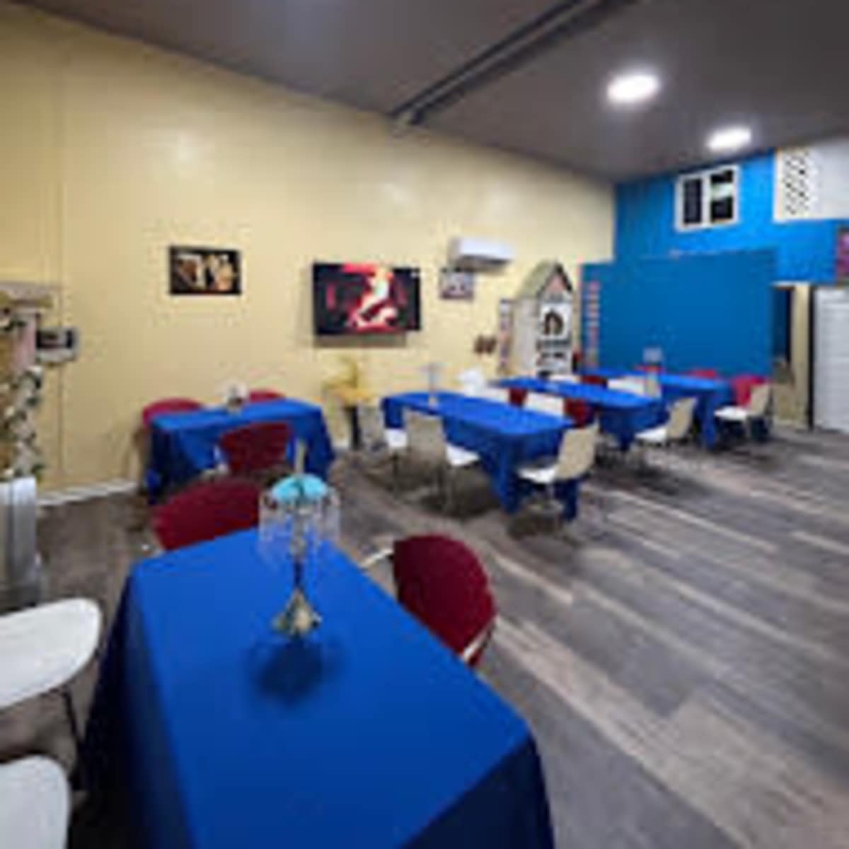 A dining area with several tables covered in blue tablecloths and red chairs, along with a television mounted on the wall displaying a fireplace scene.