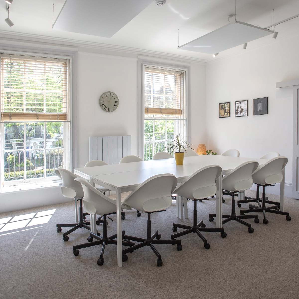 A brightly lit meeting room features a large white table surrounded by eight white chairs, with windows offering a view of greenery outside.