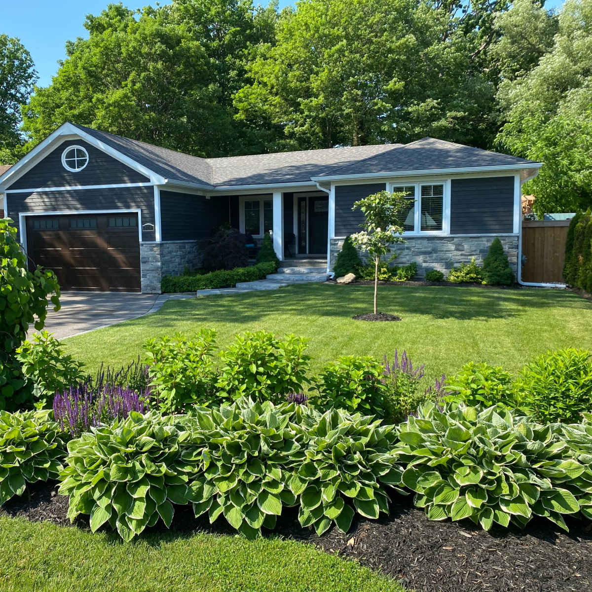 A single-story blue house with a brown garage is set in a well-kept yard featuring neatly trimmed shrubs and colorful flowers.