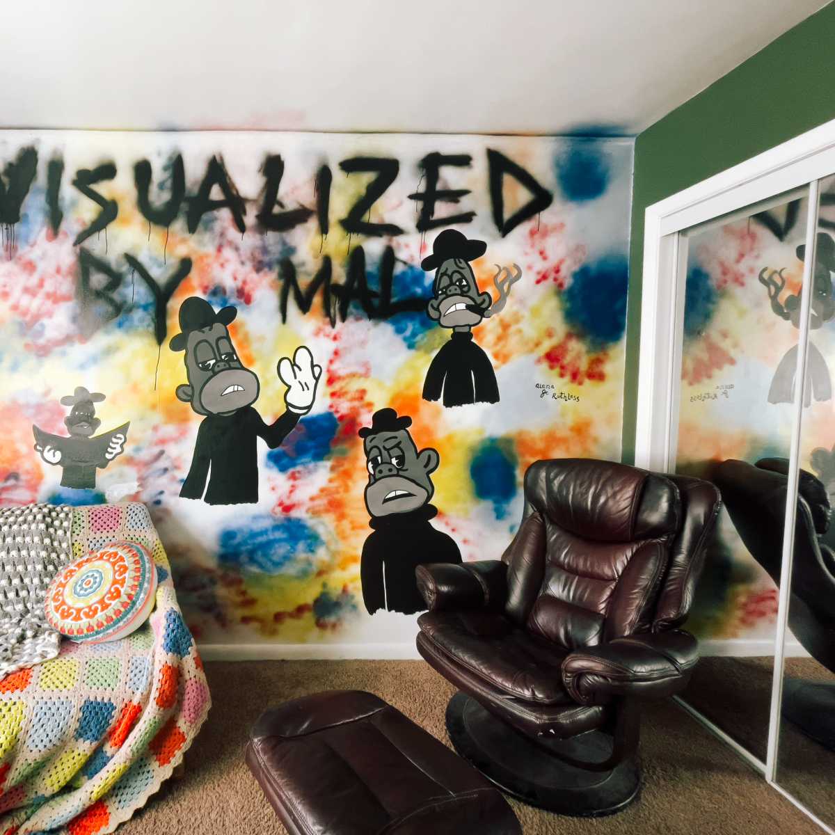 A room with a colorful graffiti mural featuring cartoon characters and the text "VISUALIZED BY MAL" on one wall, along with a patterned blanket and a brown leather chair.