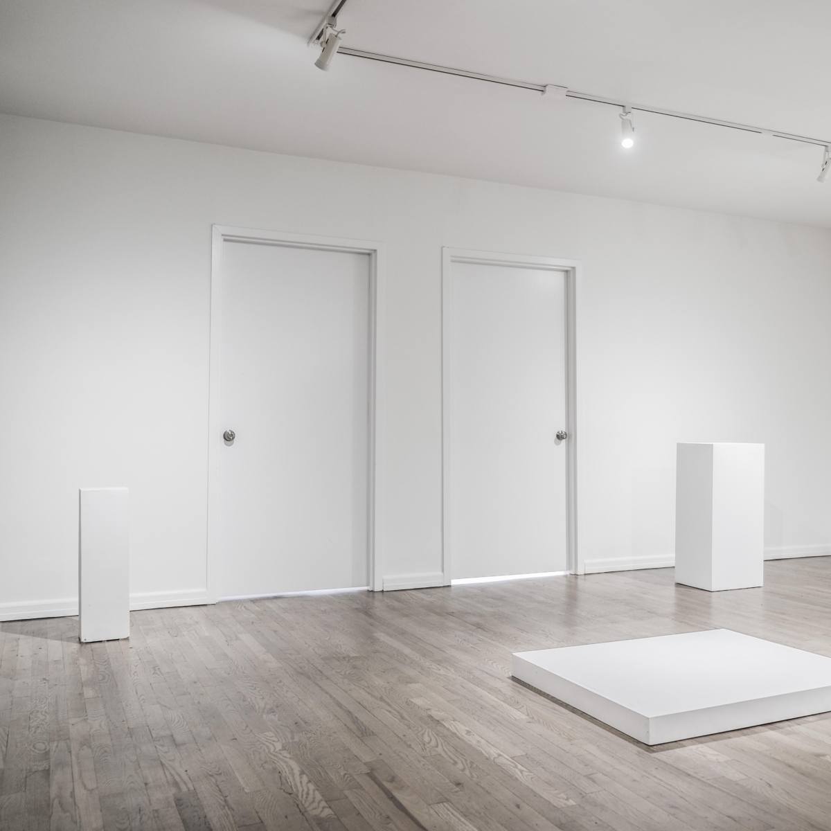 A minimalist gallery space with three white doors and several white pedestals arranged on a wooden floor.