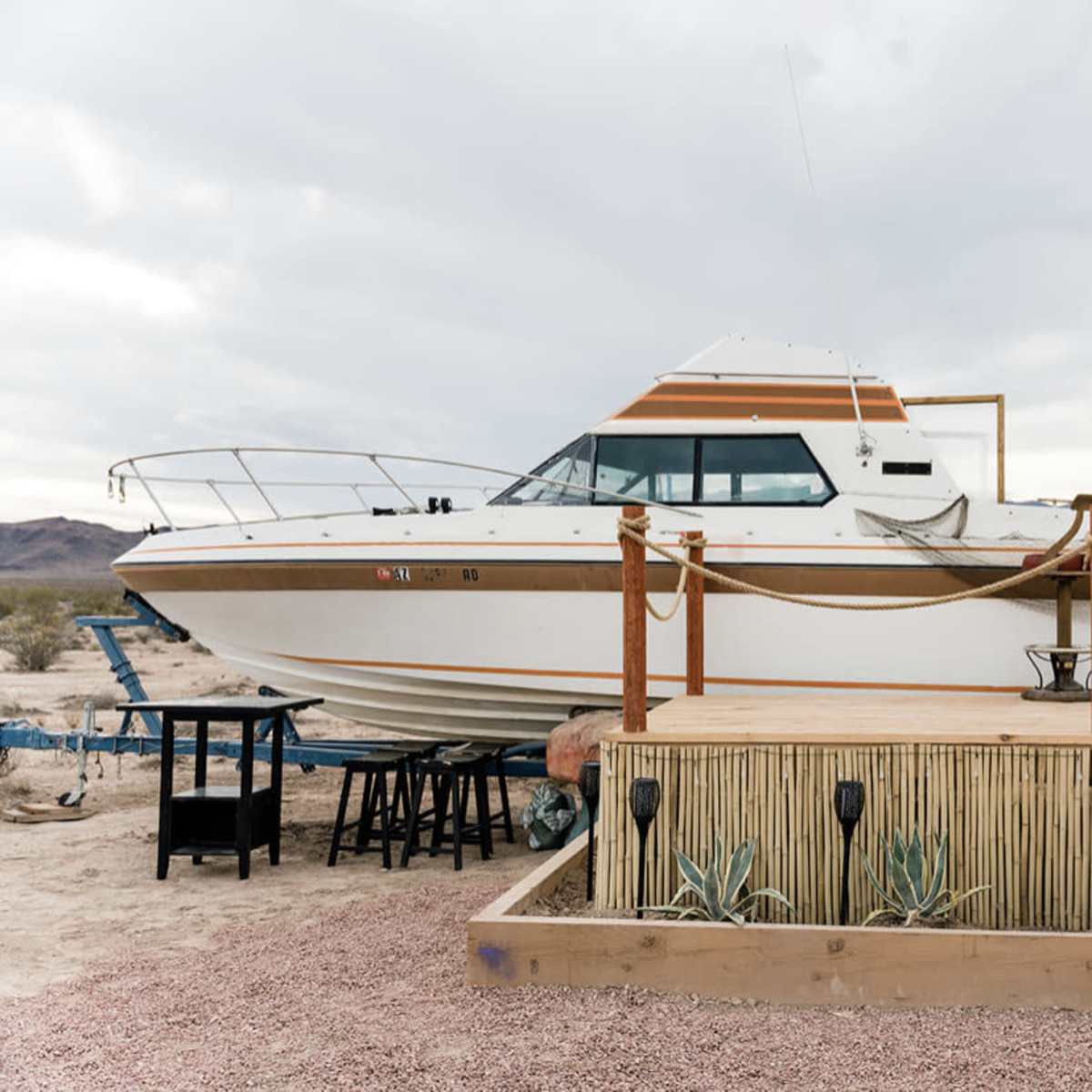 A boat is perched on a raised wooden platform in a desert landscape, with a few tables nearby and mountains visible in the background.