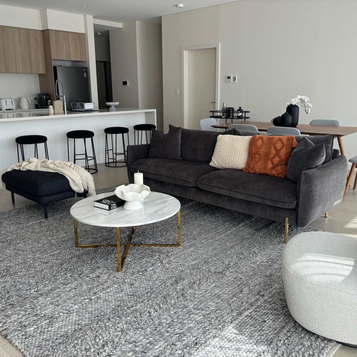 A modern living room with a gray couch, a round coffee table, and a light-colored area rug, along with a kitchen area in the background.