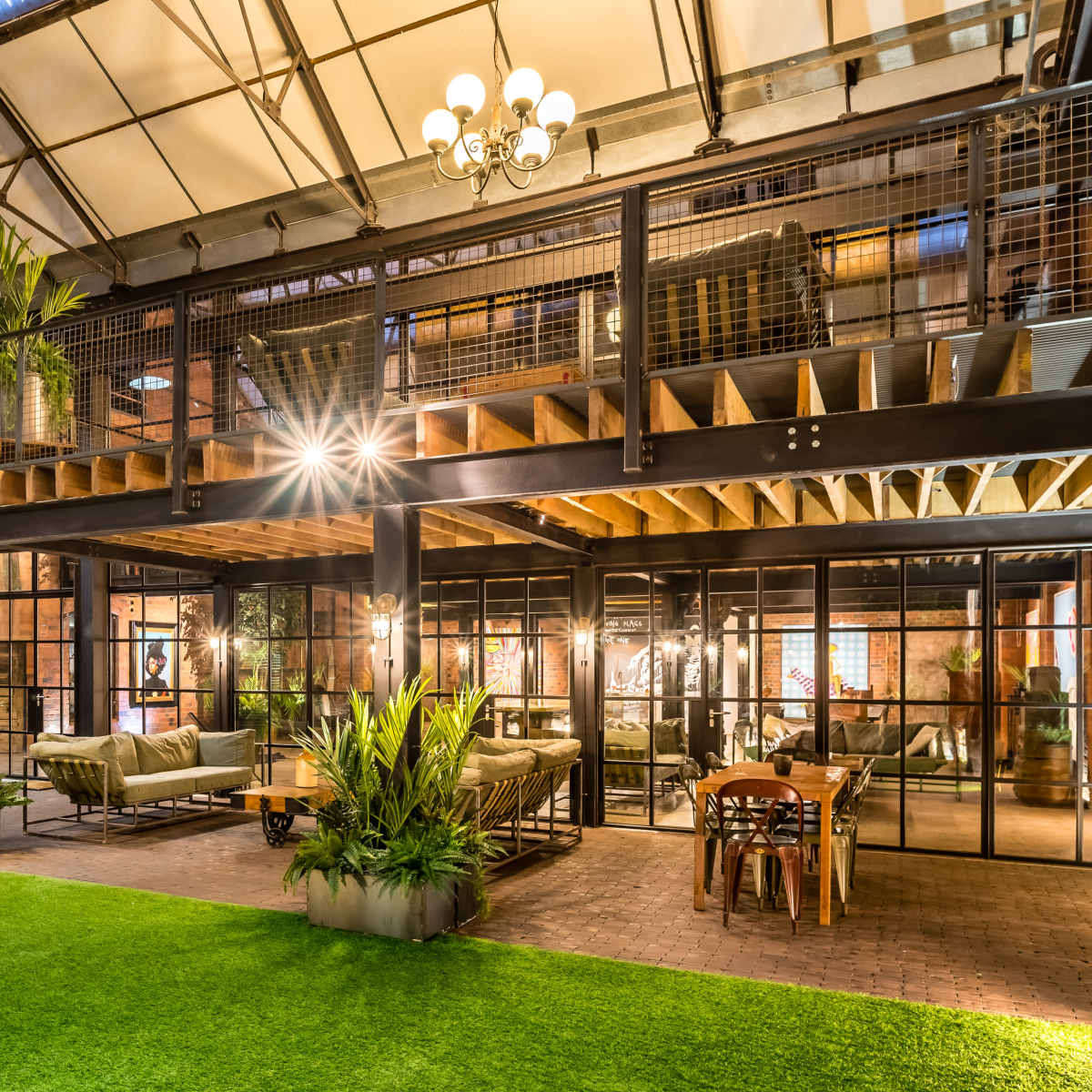 An industrial-style interior space featuring a large open area with a green lawn, plants, and metal and wooden furnishings, illuminated by multiple light fixtures.
