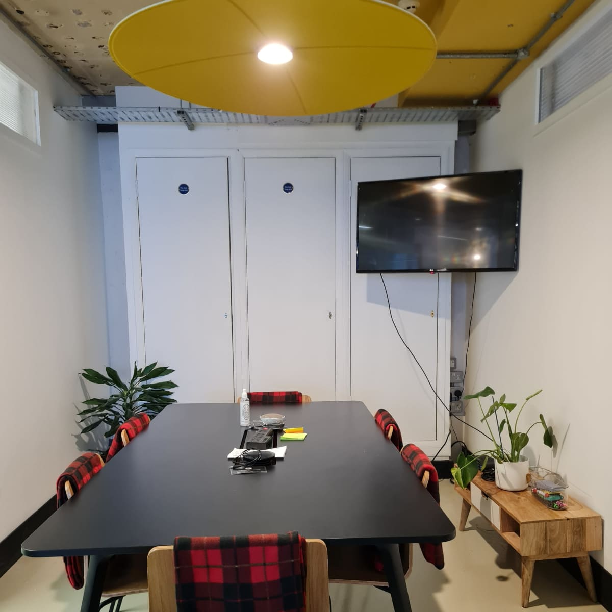 A modern meeting room features a black table surrounded by chairs with plaid seat covers, a television mounted on the wall, and plants in wooden planters.