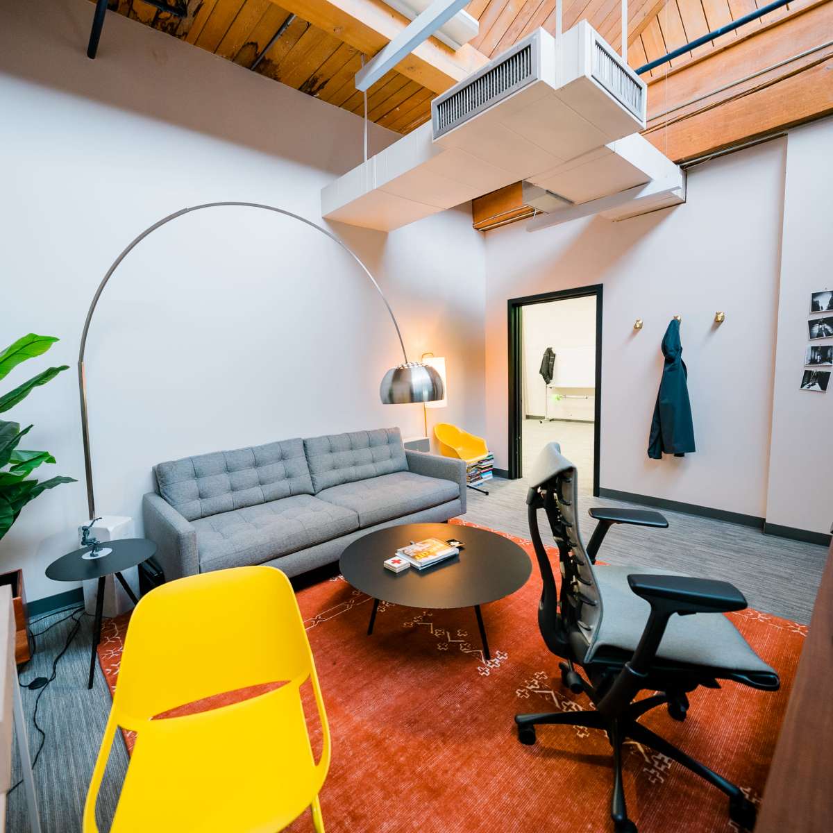 A modern office space featuring a gray sofa, a round coffee table, a desk with a computer, a potted plant, and yellow chairs on a patterned rug.