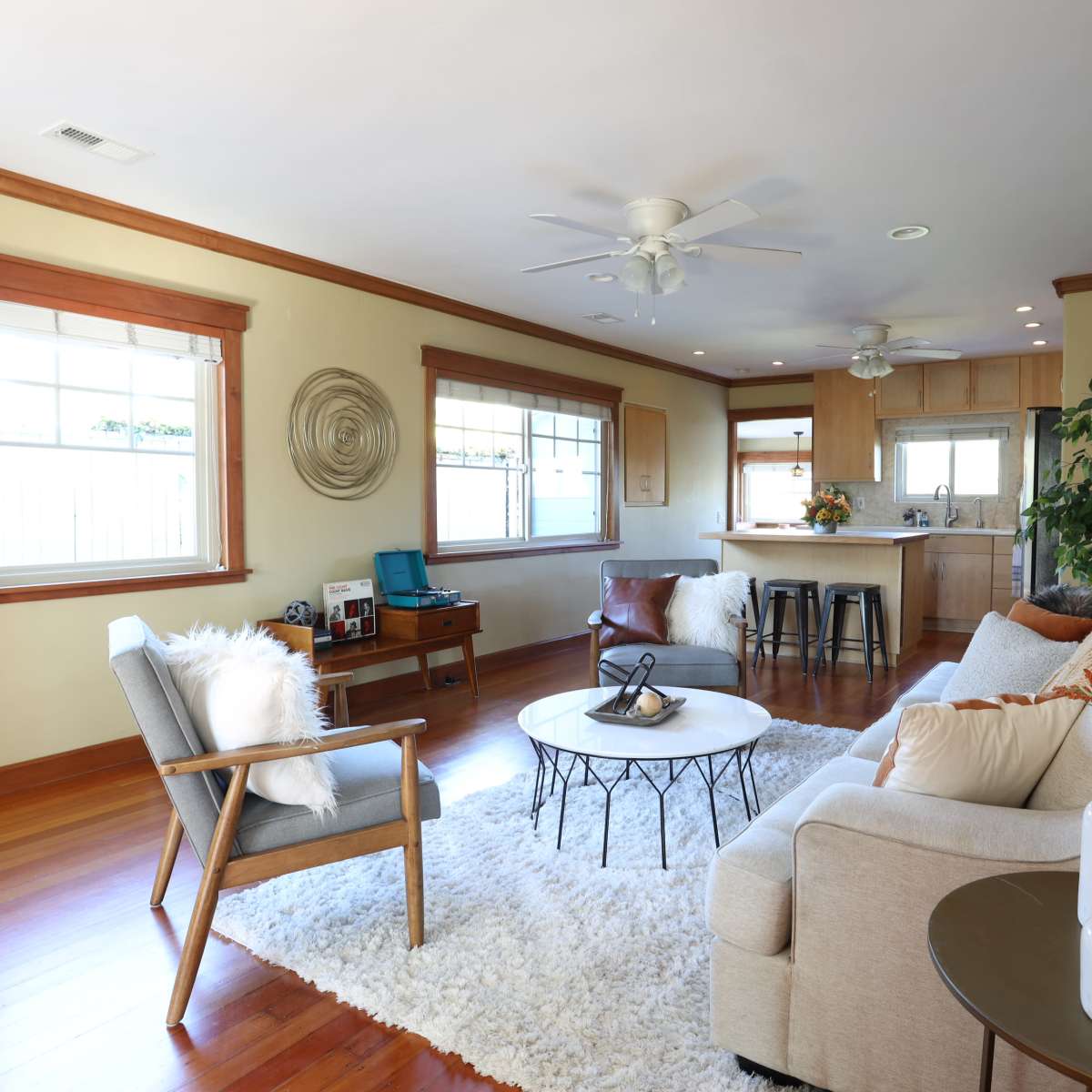 A light-filled living room features a cozy sectional sofa, a round coffee table, and a textured area rug, with a kitchen in the background.