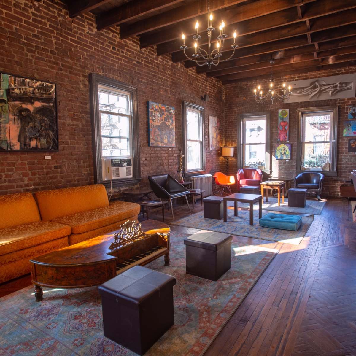 The room features exposed brick walls, a large orange sofa, various artworks, and a mix of seating arrangements, illuminated by overhead lighting.