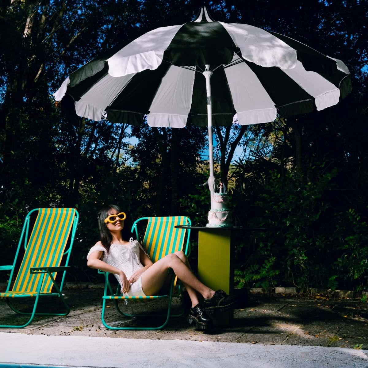 A person relaxes in a striped deck chair under a large umbrella by a pool, surrounded by greenery.