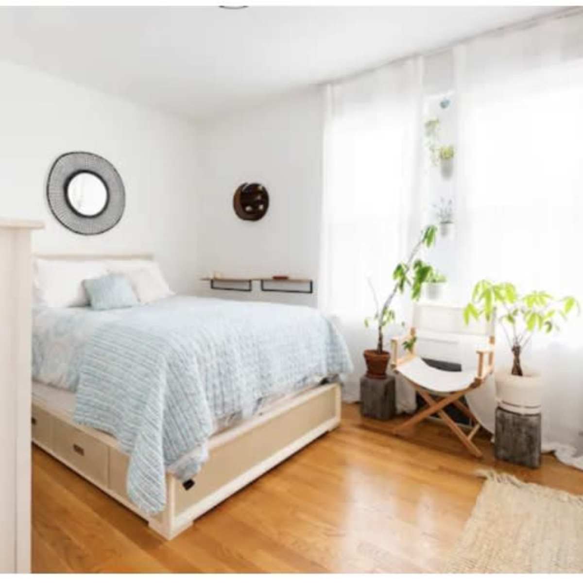 A bright bedroom with a bed, a circular mirror on the wall, a small round table, and plants in natural light.