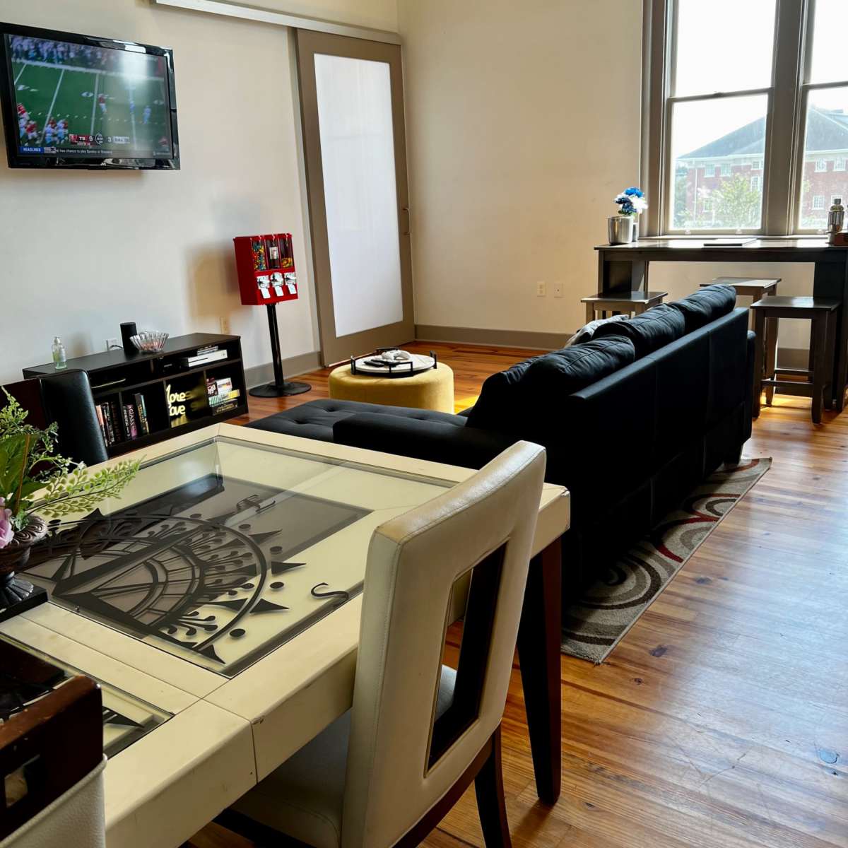 A modern living space features a dining table with a decorative design, a black sofa, a TV displaying a sports game, and a large window illuminating the room.