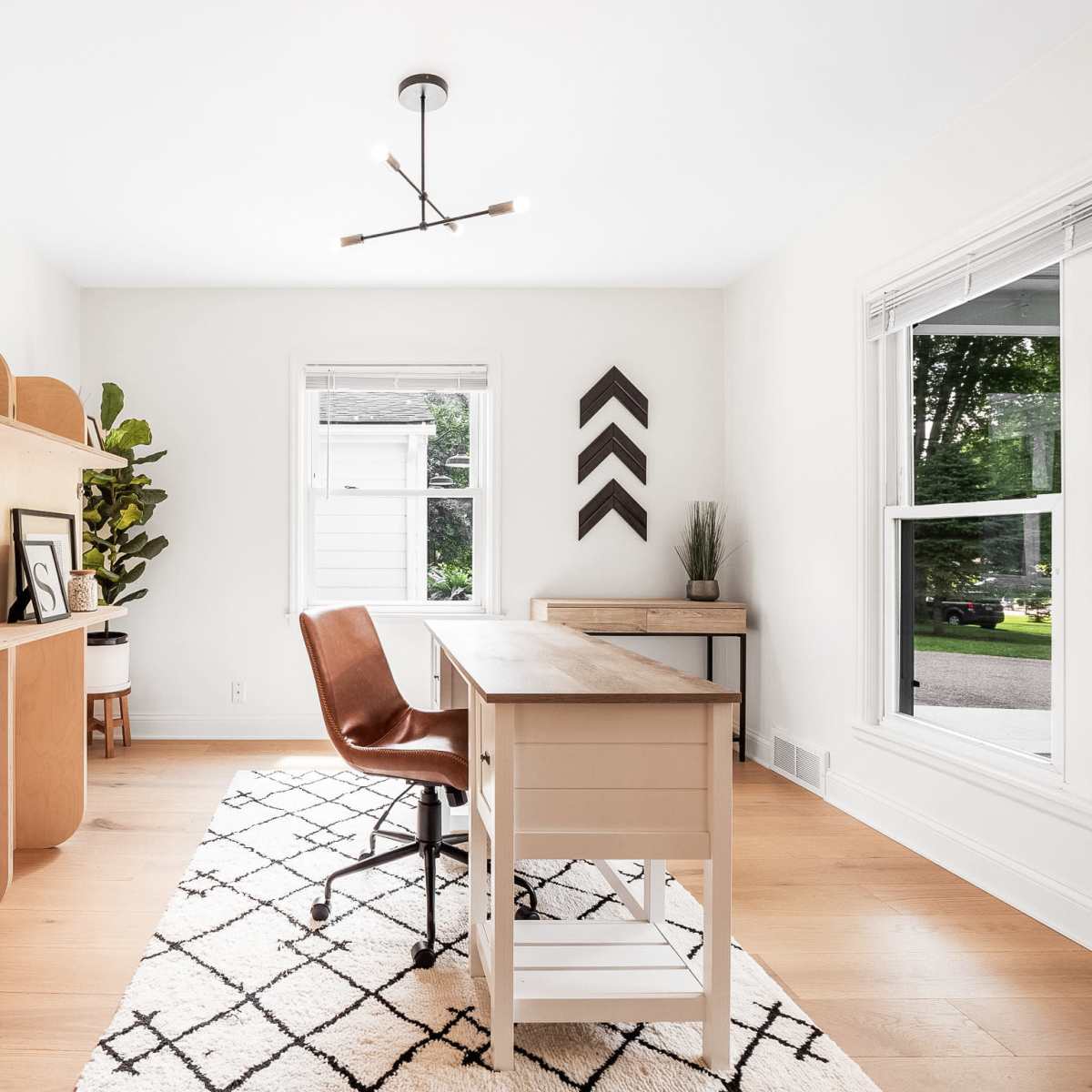 A modern home office featuring a wooden desk with a leather chair, a large shelving unit, a potted plant, and large windows allowing natural light.