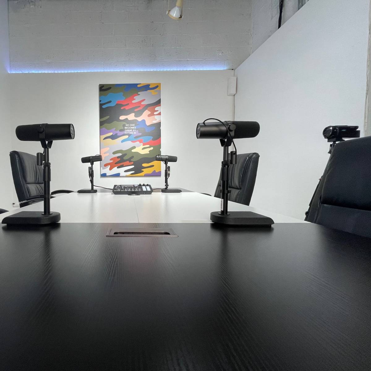 A conference room features a round table with several microphones and black leather chairs, set against a colorful abstract wall mural.