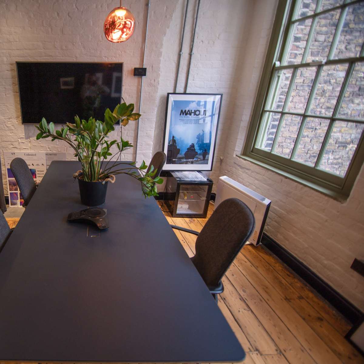A modern conference room features a rectangular black table surrounded by four chairs, with a potted plant in the center and a wall-mounted television in the background.