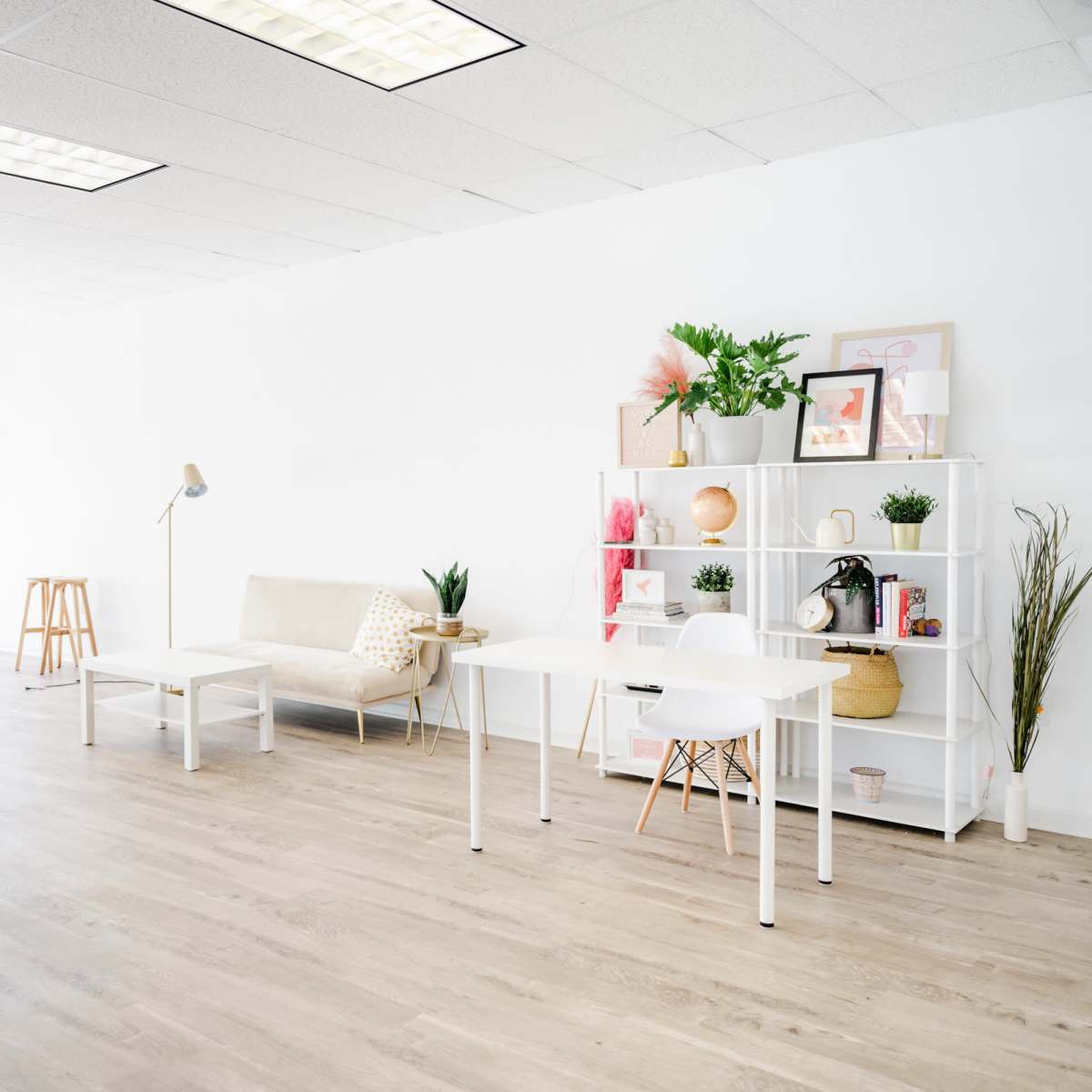 A bright, minimalistic room with light wooden flooring, featuring a beige sofa, a simple white bookshelf, and two white tables, along with natural light coming through large windows.