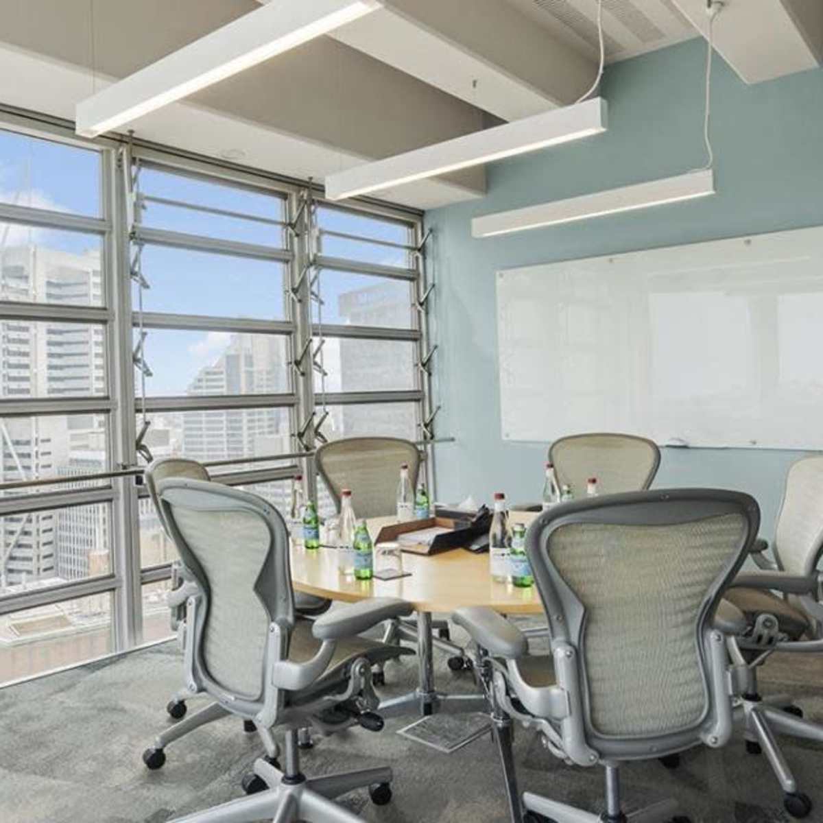 A conference room with a round table surrounded by six chairs, large windows offering a view of a city skyline, and a whiteboard on the wall.