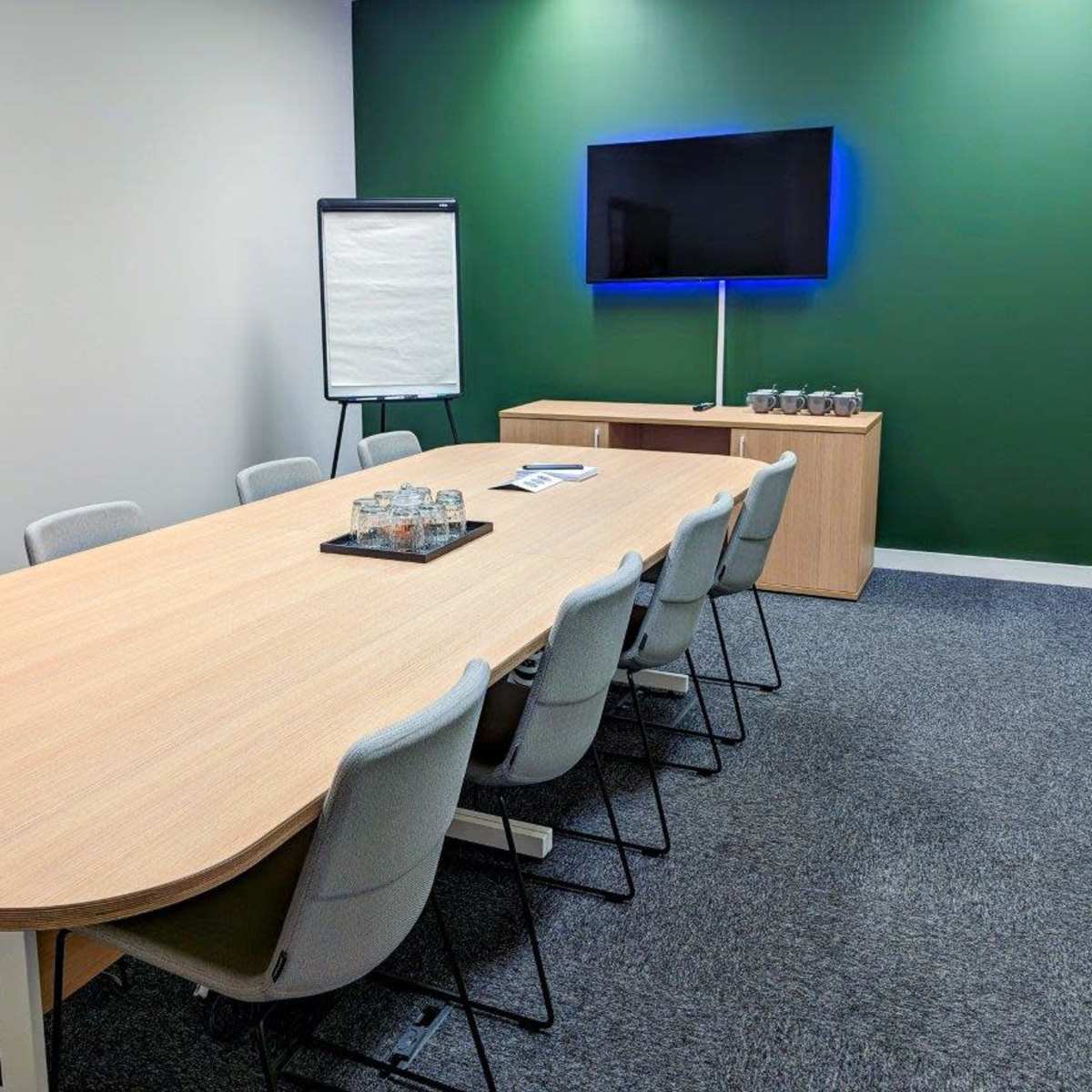 A conference room with a long wooden table, eight chairs, a whiteboard on an easel, a television mounted on a green wall, and a side cabinet displaying glassware.