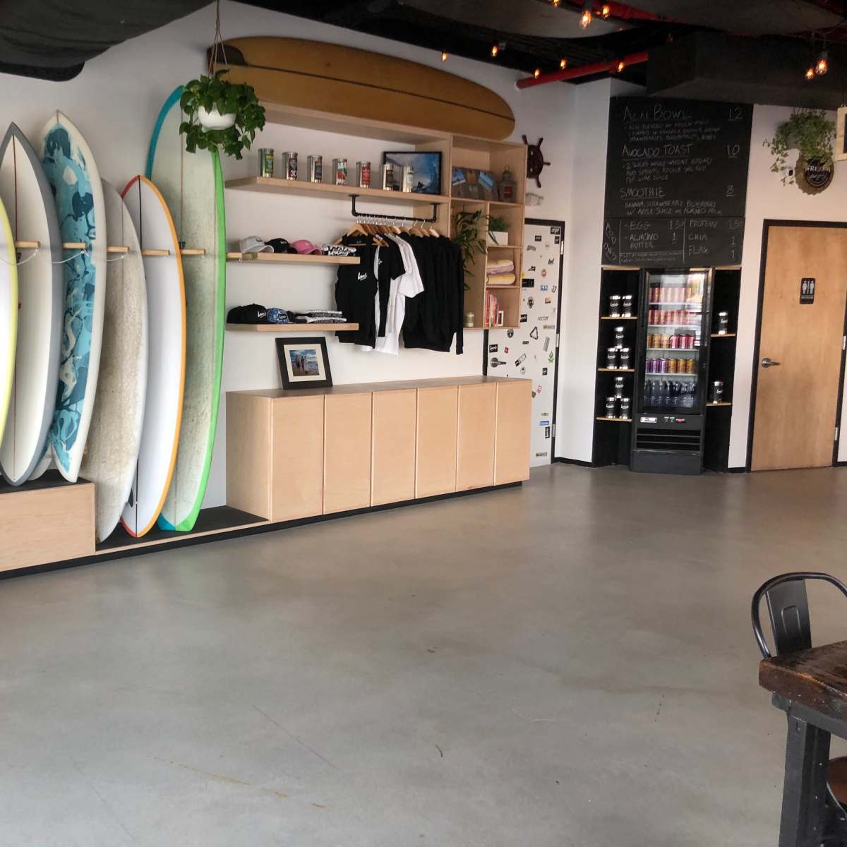 A surf shop interior with multiple surfboards displayed on a wall, clothing hanging on a rack, and a refrigerator against the back wall.