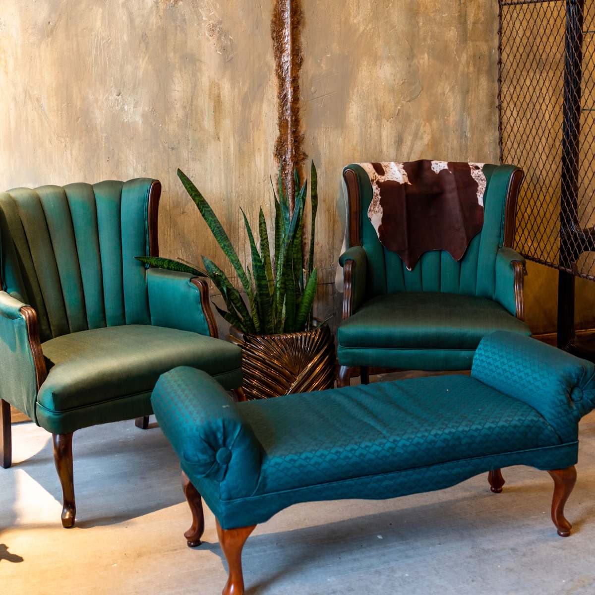 A seating area with two teal upholstered chairs and a teal chaise longue, accompanied by a large plant in a textured pot against a textured gold wall.