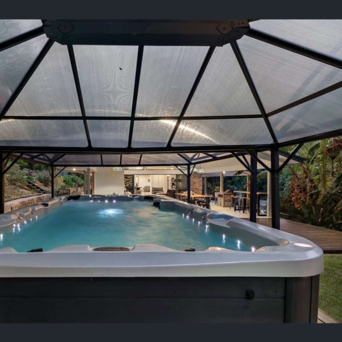 A spacious indoor swimming pool housed under a glass-roofed structure, surrounded by greenery and leading to an outdoor seating area.